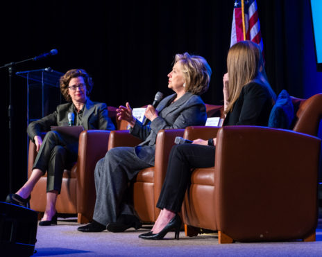 Hilary Clinton speaking on stage with two other women, all sitting in chairs